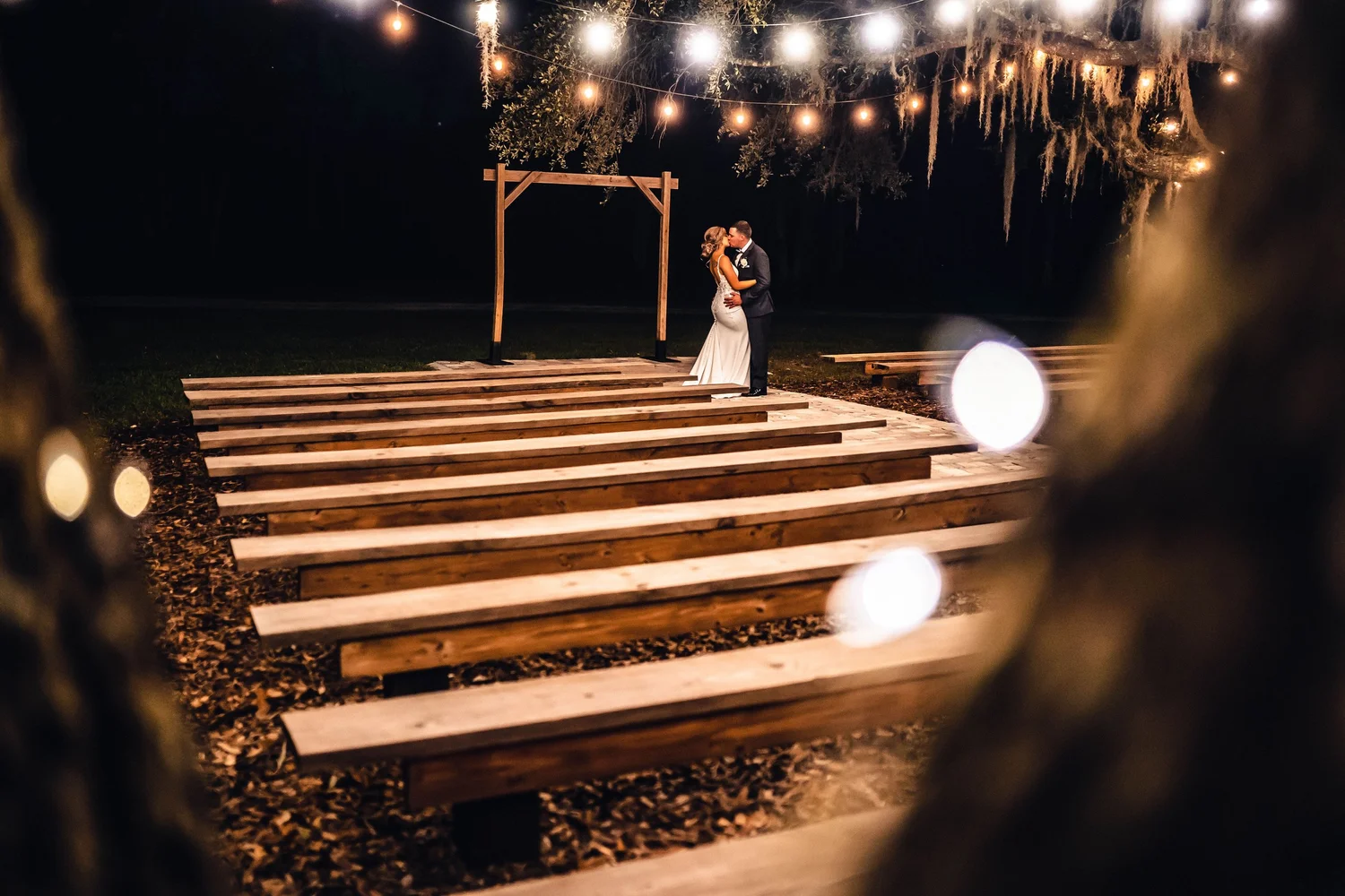 The ceremony benches lit by string lights at night, a couple under the arbor at the far end