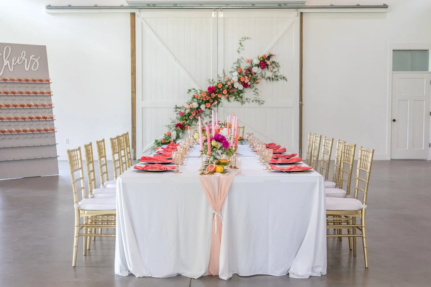 A long reception table with gold chairs and a pink floral runner inside the white barn